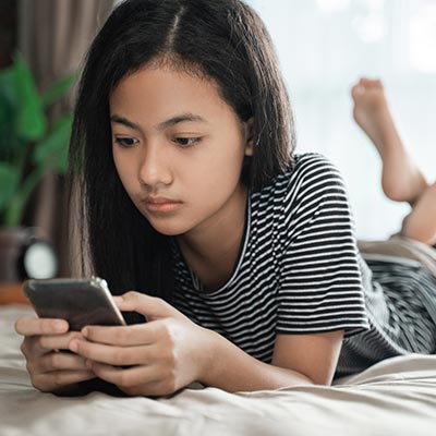 A depressed adolescent girl laying on a bed looking at her phone.